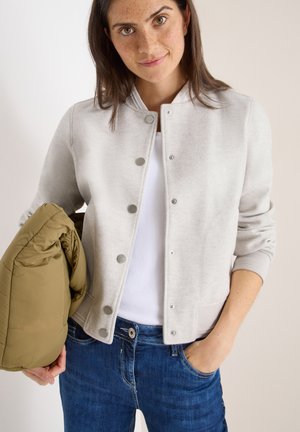 Hellgraue Bomberjacke aus glattem Stoff, mit Druckknöpfen, gerippten Bündchen und Kragen. Das Modell hält eine olivgrüne Daunenjacke.