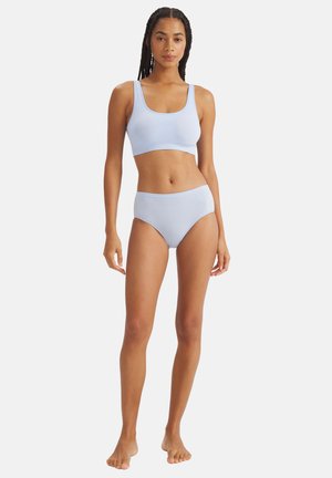 Vrouw staat blootsvoets, draagt een lichtblauwe naadloze sportbeha en hoge taille ondergoed tegen een effen witte achtergrond.