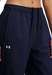 Marineblauwe sportbroek met een elastische tailleband, trekkoord, zijzakken en een zilveren logo op de linker dij. Een gladde stoftextuur.