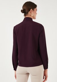 Blusa viola a maniche lunghe con colletto alto, tessuto morbido, design aderente e polsini con bottoni, che mostra una vista posteriore sagomata.