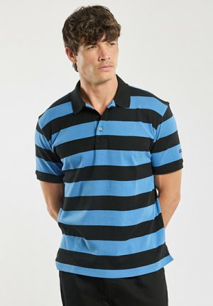 Homme portant un polo à rayures horizontales bleues et noires, debout les mains derrière le dos, regardant sur le côté sur un fond blanc.