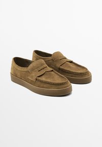 Braune Wildleder-Schlupfschuhe mit gesticktem Design und einem dekorativen Riemen auf der Vorderseite, auf einem weißen Hintergrund dargestellt.