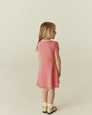 Νεαρή κοπέλα με μακριά ξανθά μαλλιά, που κοιτάζει πλάγια, φορώντας ένα ροζ φόρεμα, λευκές κάλτσες με βολάν και κρεμ παπούτσια, με φόντο ένα απλό υπόβαθρο.