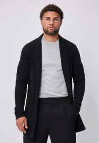Homme aux cheveux bouclés et à la barbe, portant une chemise gris clair, un cardigan noir ouvert et un pantalon noir, debout avec une main dans la poche.
