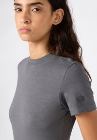 Jonge vrouw met lang, donker haar die een nauwsluitend grijs T-shirt met korte mouwen draagt, met een klein logo op de linkersleeve.