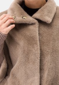 Abrigo de piel sintética marrón con cuello solapado, dos botones decorativos y textura suave. Manga de suéter de punto visible a la izquierda.