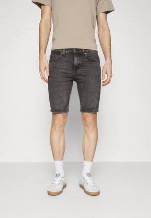 Uomo che indossa pantaloncini di jeans neri, t-shirt beige, calzini bianchi e scarpe da ginnastica bianche, in piedi davanti a uno sfondo chiaro e semplice.