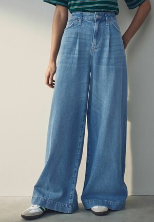 Personne portant un jean bleu taille haute à jambes larges, une chemise rayée verte et noire, et des baskets blanches avec des rayures vertes, debout contre un mur uni.