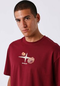 Hombre con cabello corto, vistiendo aretes de aro plateados y una camisa burdeos con diseño floral y texto "PXP", mirando a la cámara frente a un fondo llano.