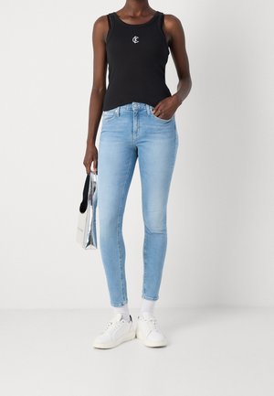 Personne portant un jean skinny bleu clair, un top noir sans manches avec un logo, des baskets blanches, des chaussettes blanches et tenant un sac cabas argenté.