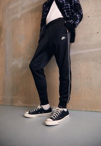 Osoba oblečená v černých Nike teplácích s bílým pruhem a černých nízkých teniskách stojí proti texturované béžové zdi.