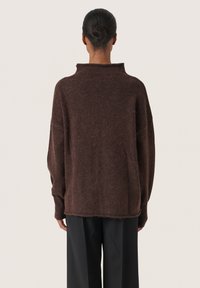 Braun, übergroßer Wollpullover mit hohem Kragen, fallenden Schultern und rohem Saum. Kombiniert mit schwarzen, weiten Hose.