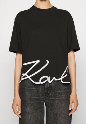 Person wearing a black short-sleeve t-shirt with "Karl" in large white script across the lower front, paired with dark gray jeans.