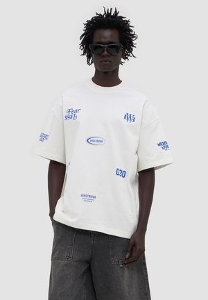 Personne à la peau foncée et aux cheveux courts bouclés, portant des lunettes de soleil noires, un t-shirt blanc oversize avec un texte bleu, et un pantalon gris foncé.