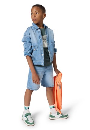 Denim jacket, green graphic T-shirt, light blue denim shorts, orange sweatshirt in hand, white and green sneakers with striped socks.