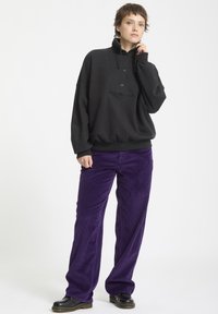 Felpa nera con colletto alto e bottoni frontali, abbinata a pantaloni a gamba larga in corda violacea. Scarpe nere completano l'outfit.