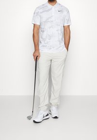 Muž v světlém golfovém oblečení, držící golfovou hůl, stojící na jednoduchém bílém pozadí.