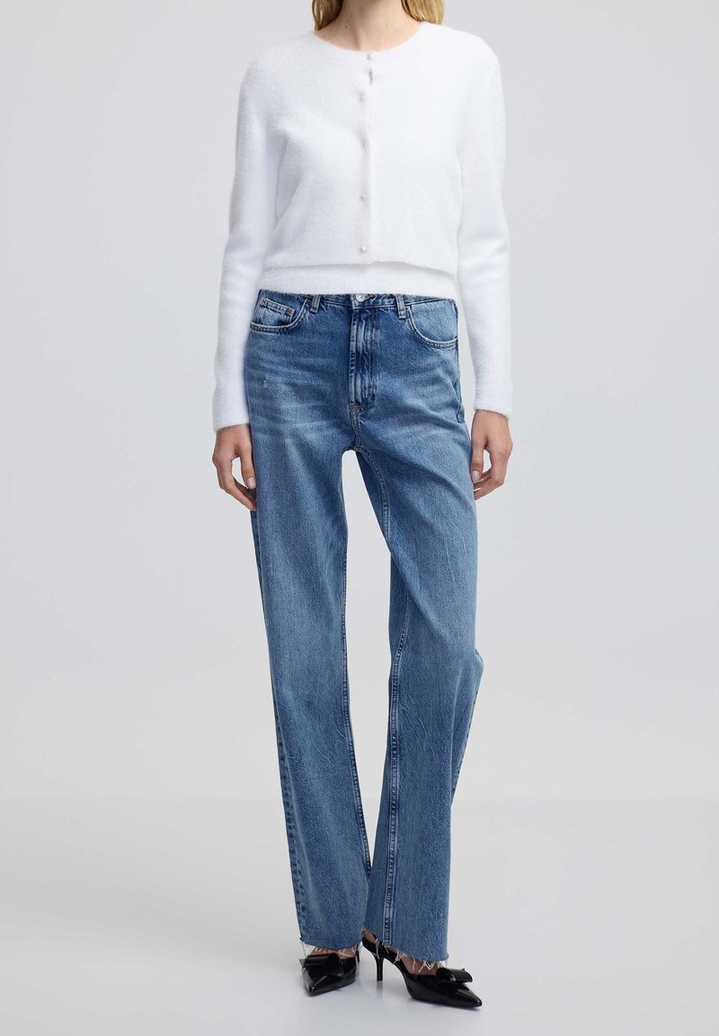 Femme portant un cardigan court blanc à boutons en tissu duveteux, un jean bleu taille haute droit, et des chaussures noires à talons aiguilles pointues avec des nœuds.