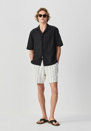 Fiatal férfi fekete rövid ujjú, gombos ingben, fehér csíkos rövidnadrágban, fekete papucsban és napszemüvegben, zsebre tett kézzel áll.