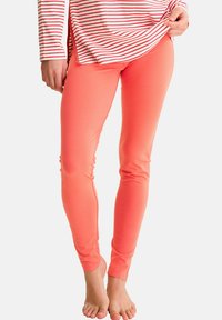 Femme portant des leggings corail ajustés et un t-shirt à manches longues rouge et blanc rayé, levant légèrement le t-shirt, pieds nus sur un fond blanc.