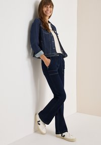 Giacca in denim scuro con maniche arrotolate, top crema chiaro, jeans a zampa scuri e sneakers bianche con logo nero, appoggiata a un muro.