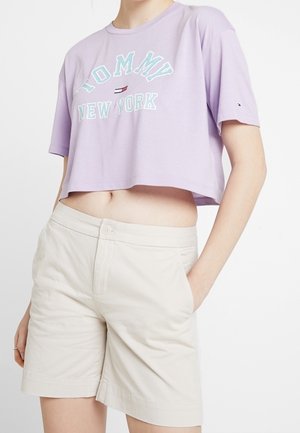 Person iført en lavendel kort t-shirt med teksten "Tommy New York" og beige højtaljede shorts med den ene hånd i lommen.