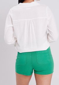 Chemisier blanc à manches longues avec un détail de empiècement au dos, associé à un short en denim vert avec poches et une coupe ajustée.