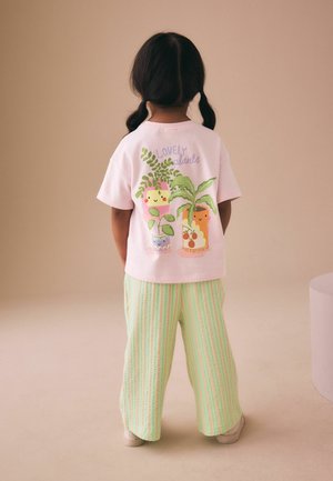 Kind mit dunklem Haar in Zöpfen, das ein rosa T-Shirt mit Cartoon-Pflanzen und gestreifte grüne Hosen trägt, steht vor einem neutralen Hintergrund.