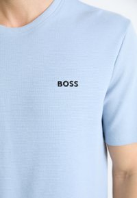 Svetlo modré tričko s krátkym rukávom vyrobené z textúrovanej látky, s čiernym logom "BOSS" na ľavej strane hrude.