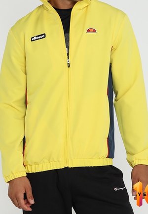 Osoba ubrana w żółtą kurtkę zapinaną na zamek błyskawiczny z logo ellesse na piersi oraz czarne spodnie z logo Champion na udzie.