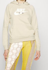 Beige hoodie med vit "NIKE" logotyp, med känguruficka och dragsnören. Matchad med mönstrade multikolor leggings.
