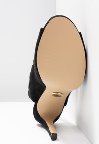 Czarny zamszowy but na wysokim obcasie ustawiony pod kątem, ukazujący gładką beżową podeszwę i wąski czubek obcasa na białym tle.