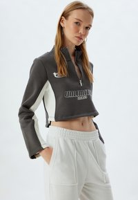 Sudadera corta gris y blanca con cuello con cremallera, mangas largas y el texto "ENERGÍA ILIMITADA". Combinada con pantalones blancos de cintura elasticada.