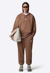 Brun hoodie och sweatpants med en vit logotyp, parat med grå sneakers och en krämfärgad tote bag. Solglasögon fulländar outfiten.