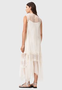 Durchscheinendes cremefarbenes Kleid mit einem geschichteten Spitzen-Saum, ausgestattet mit einem strukturierten Netzoberteil und einem High-Low-Design. Betont durch ein Rückendetail mit Schlüsselloch-Ausschnitt.