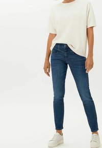 Camisa blanca de manga corta combinada con jeans ajustados de color azul oscuro y zapatillas blancas. Los jeans tienen un bolsillo en la parte frontal y un detalle de botón metálico.