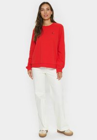 Roter Pullover mit langen Ärmeln, rundem Ausschnitt und kleinem schwarz gesticktem Detail auf der Brust. Kombiniert mit weißen weiten Hosen und Turnschuhen.