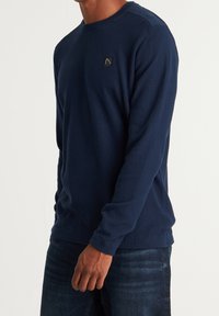 Marinblå stickad tröja med långa ärmar och ribbade muddar, med ett strukturerat mönster och en liten logopatch på bröstet.