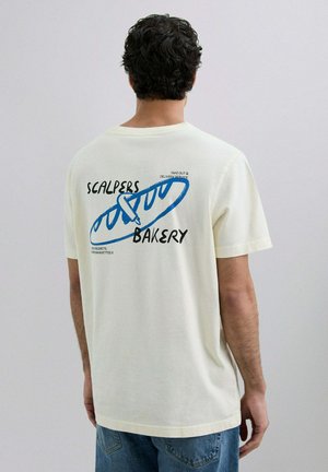 Camiseta de algodón color crema con mangas cortas, que presenta un diseño gráfico en azul y negro de una baguette y texto en la parte posterior.