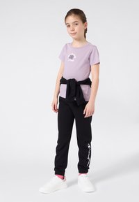 Jauna mergaitė stovi su juodu džemperiu, surištu aplink liemenį, vilkėdama alyvinį marškinėlį su užrašu "Cuties Club", juodas kelnes, rožines kojines ir baltus sportbačius.
