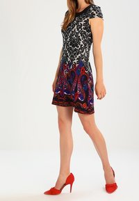 Robe à manches courtes avec un motif floral en noir et blanc sur le corsage et un motif paisley coloré sur la jupe ; assortie à des escarpins rouges.