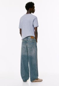 Giovane uomo di spalle, con una maglietta grigio chiaro, jeans a gamba larga di un blu sbiadito e sneakers beige su uno sfondo bianco.