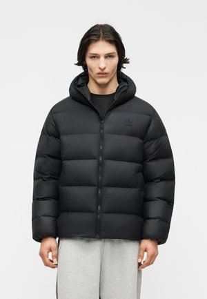 Giovane uomo con capelli scuri di media lunghezza che indossa una giacca a piumino nera con cappuccio e pantaloni della tuta grigio chiaro su sfondo neutro.