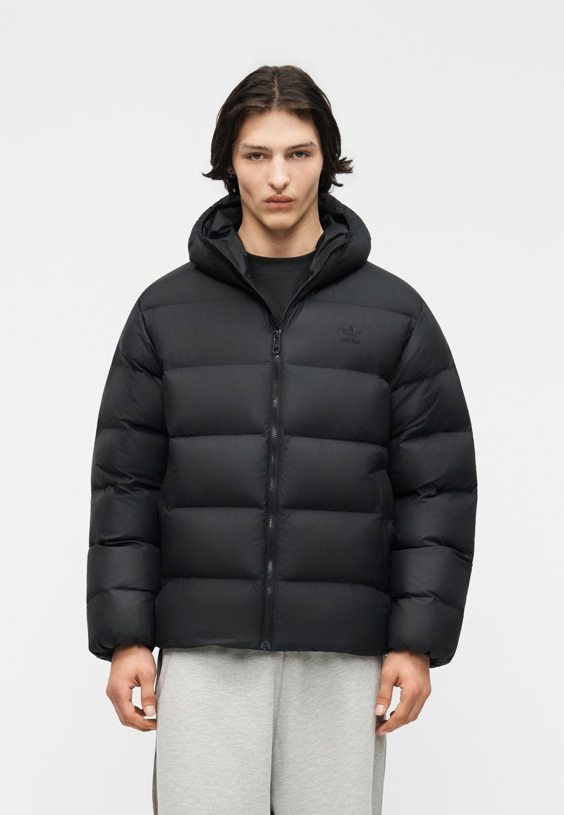 Jonge man met halflang donker haar, gekleed in een zwarte donsjas met capuchon en lichtgrijze joggingsbroek, tegen een neutrale achtergrond.