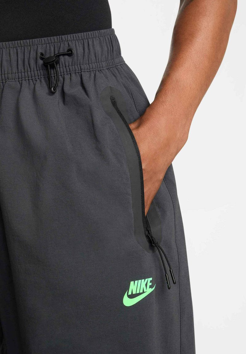 Pantalones de chándal Nike en tejido gris oscuro con cintura elástica, cordón ajustable, bolsillos laterales y un logo en verde neón en la parte frontal.