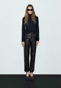 Schwarzer Rollkragenpullover, hochtaillierte schwarze Lederhose und graue spitz zulaufende Schuhe. Gerader Schnitt, minimalistisches Design, strukturierter Stoff.