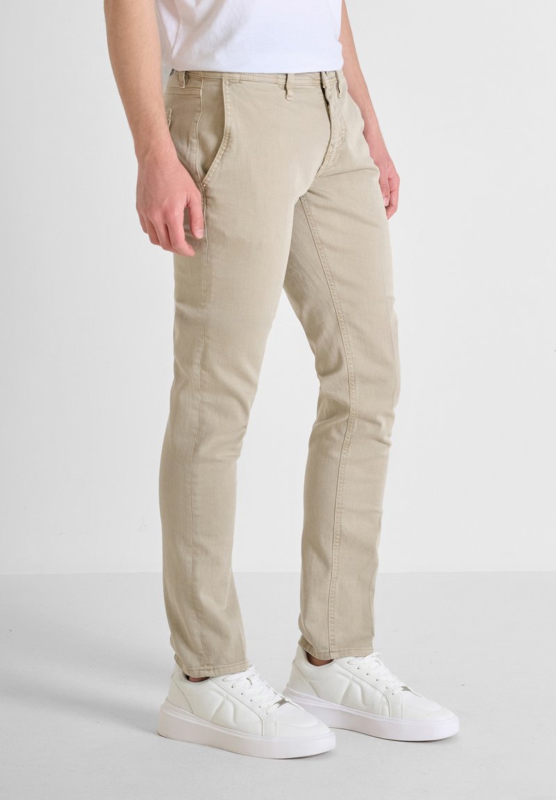 Pantalones de algodón beige con corte ajustado, con bolsillos frontales y cremallera, combinados con zapatillas blancas sobre un fondo neutro.