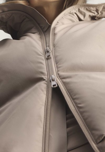Nahaufnahme einer beigen gesteppte Jacke mit teilweise geöffnetem Doppelreißverschluss, die die Textur und Nähte des Kleidungsstücks zeigt.