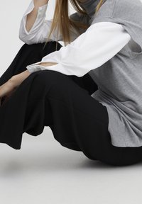 Šedý bezrukávový svetr přes bílou košili, v kombinaci s širokými černými kalhotami. Mezi rysy patří vrstvové rukávy a design s vysokým límcem.