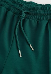 Grüne Jogginghose mit Kordelzug, elastischem Bund, glatter Textur und metallischen Spitzen an den Kordeln.